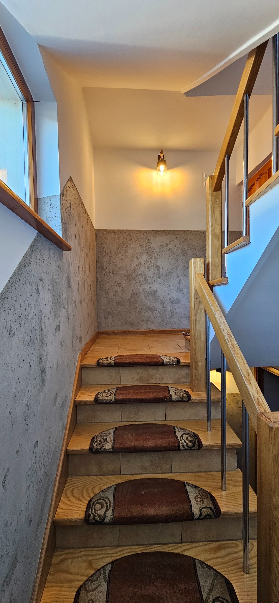 Klatka schodowa z drewnianymi schodami i metalową balustradą, wyłożona brązowymi dywanikami z ozdobnym wzorem, ściany wykończone w dwóch odcieniach szarości, oświetlenie punktowe.
