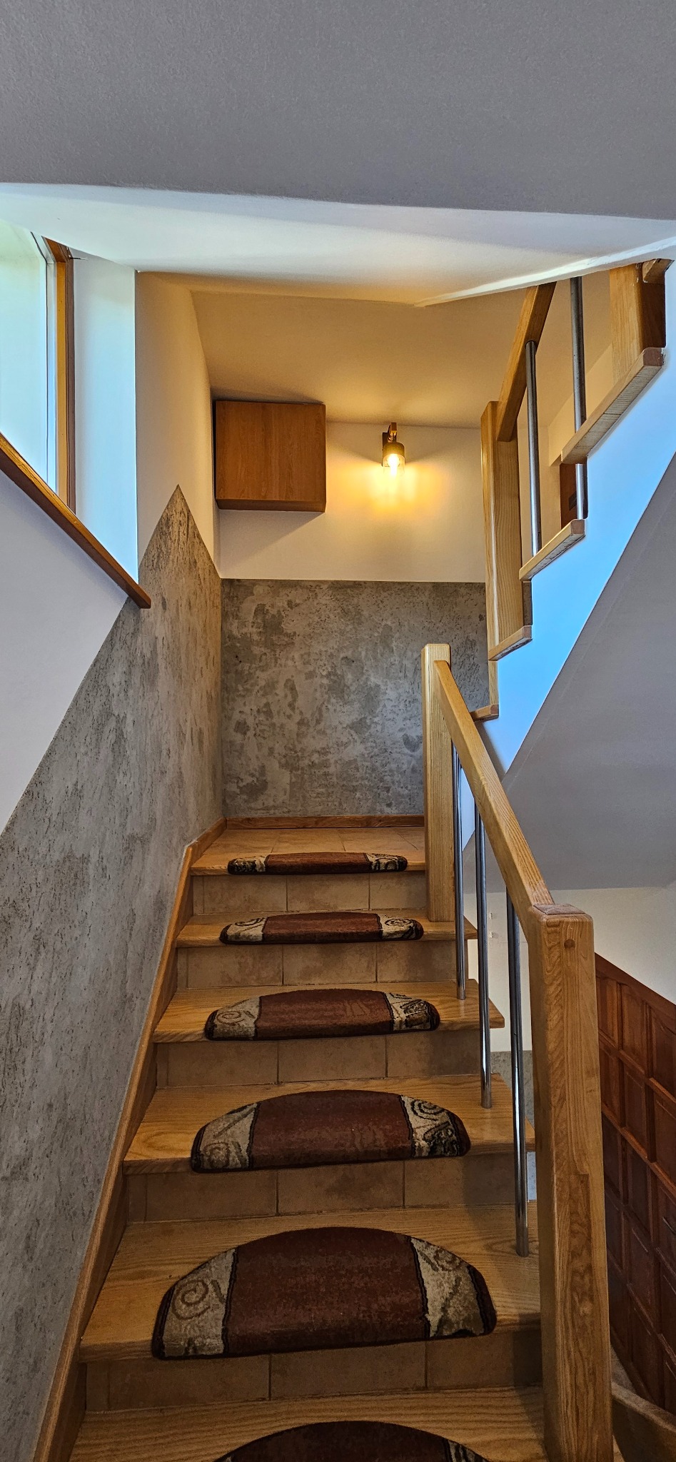 Drewniane schody z brązowymi nakładkami dywanowymi, betonowa ściana pomalowana w geometryczny wzór, drewniana szafka na ścianie i oświetlenie punktowe.