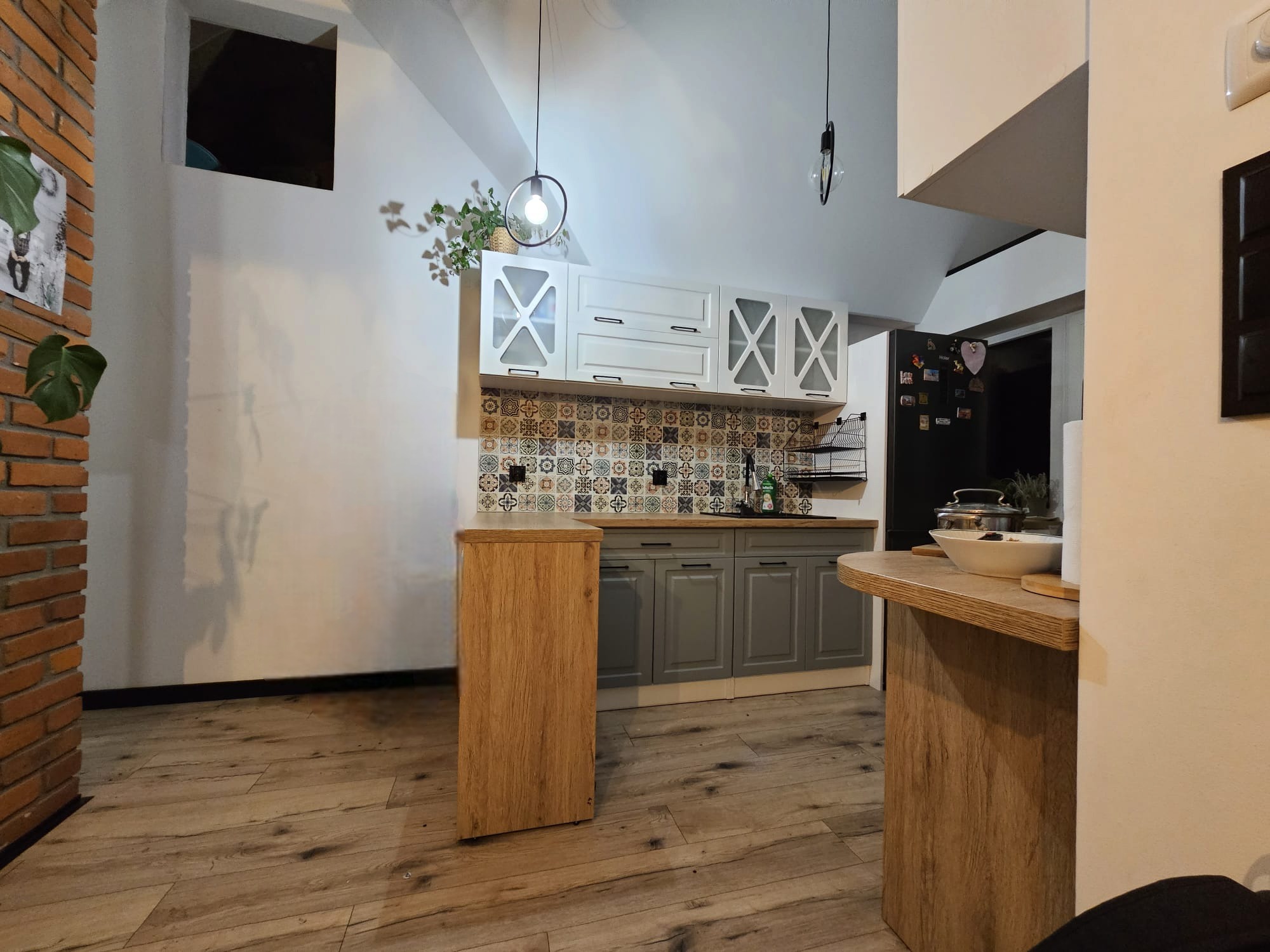Nowoczesna kuchnia z drewnianą podłogą, szarymi szafkami dolnymi i białymi górnymi z przeszklonymi frontami, ozdobiona mozaiką nad blatem i wiszącymi lampami z żarówkami Edisona.