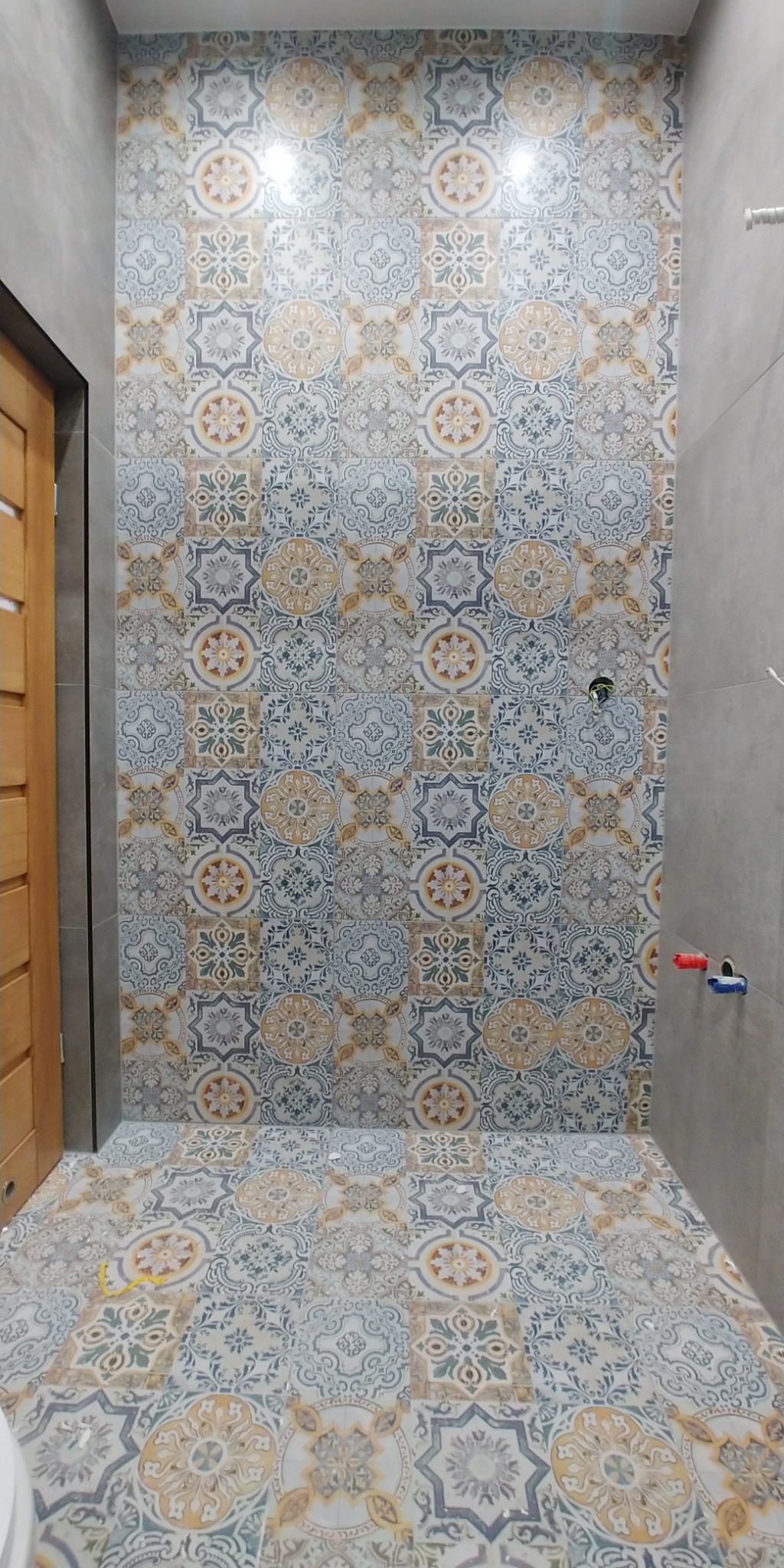 Wykończona łazienka z dekoracyjnymi płytkami podłogowymi i ściennymi w odcieniach błękitu, beżu i szarości, z widocznymi przyłączami wodnymi na ścianie i fragmentem drewnianych drzwi po lewej stronie.