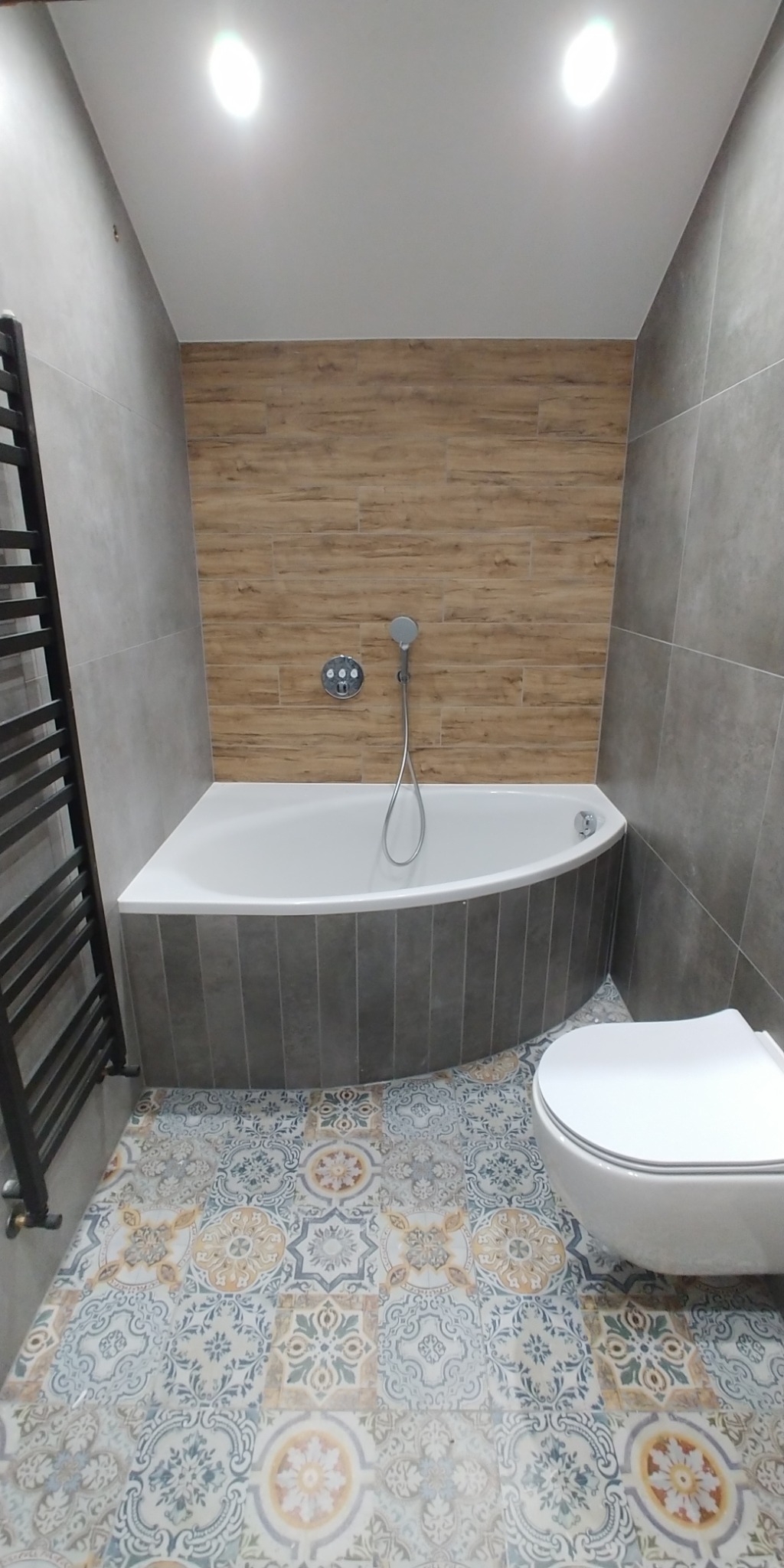 Nowoczesna łazienka z narożną wanną obudowaną szarymi płytkami, drewnopodobnymi płytkami na ścianie, mozaikową podłogą i wiszącą toaletą.