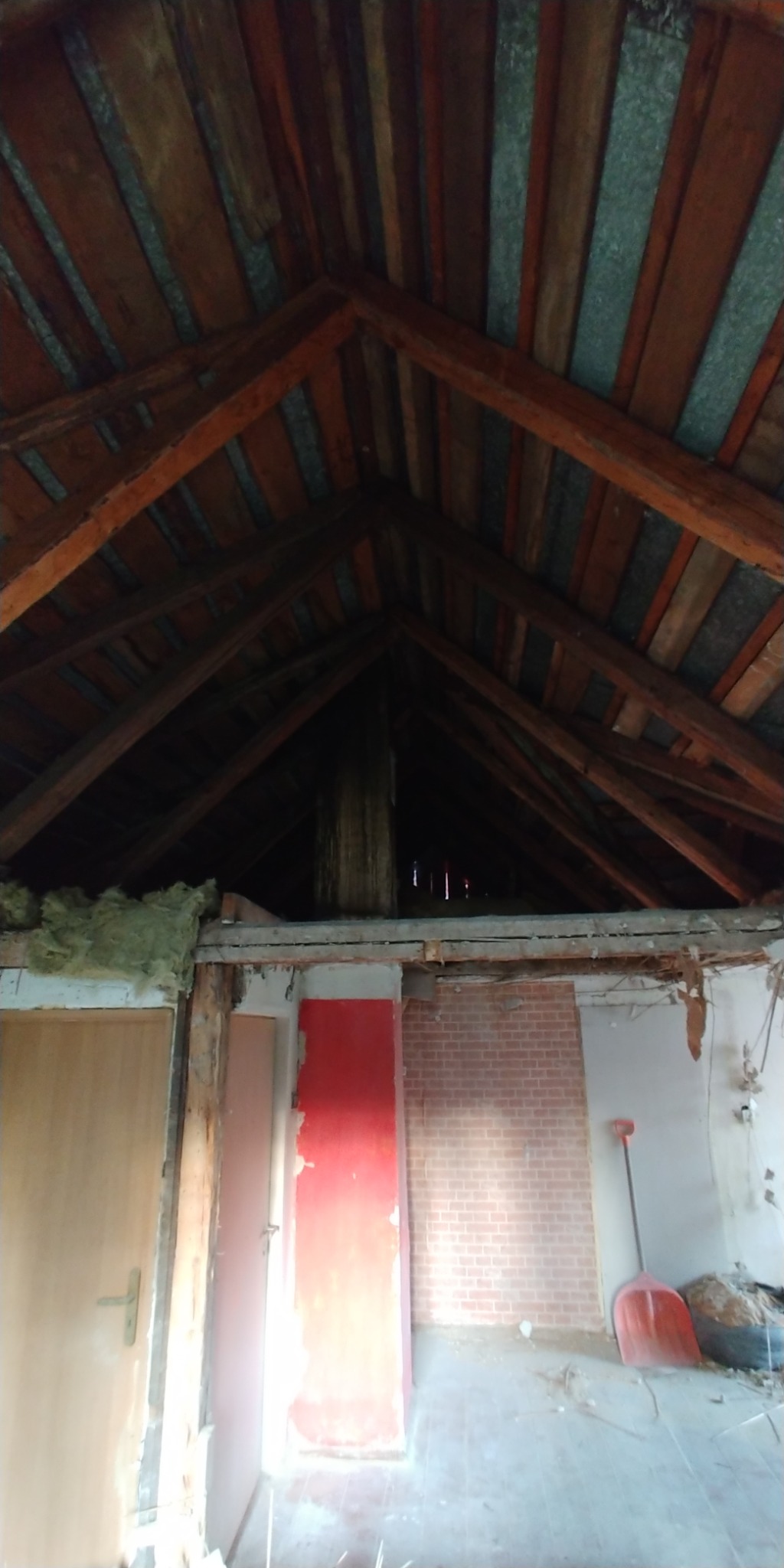 Strych w trakcie remontu: widoczne drewniane elementy konstrukcyjne dachu, fragment ocieplenia, czerwone drzwi, ceglana ściana i narzędzia.