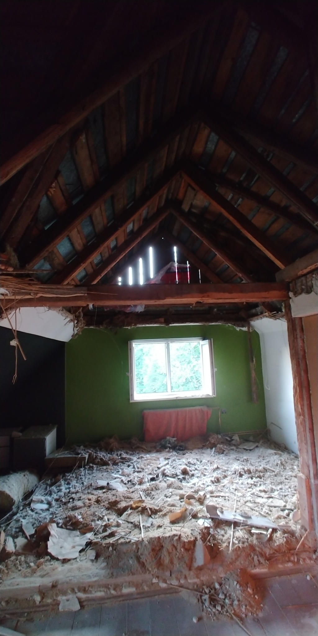 Zdjęcie poddasza w trakcie demontażu, widoczne odsłonięte krokwie dachu z prześwitami, fragmenty ścian w kolorze zielonym i białym, gruz i resztki materiałów budowlanych na podłodze.
