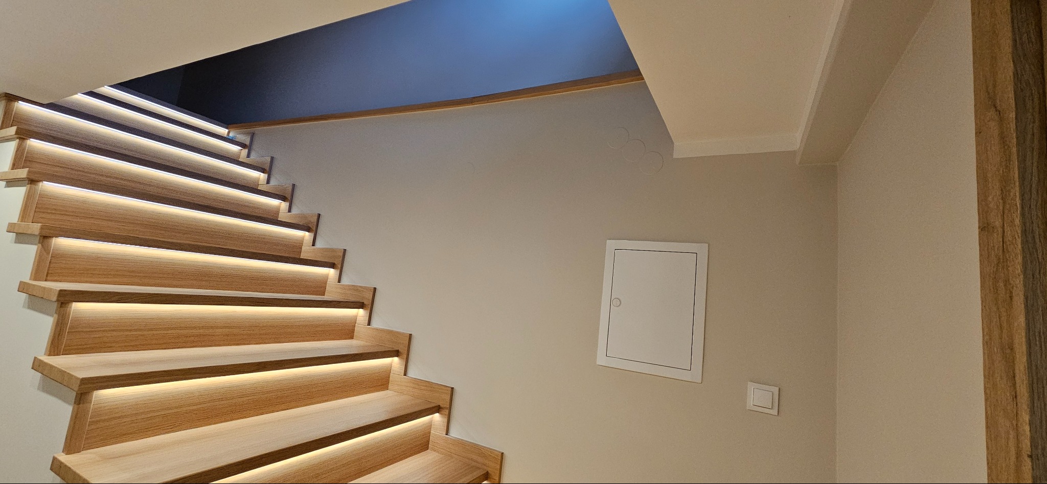 Nowoczesne drewniane schody z podświetleniem LED wzdłuż stopni, jasna ściana z drzwiczkami rewizyjnymi i włącznikiem, ciemny sufit w tle.