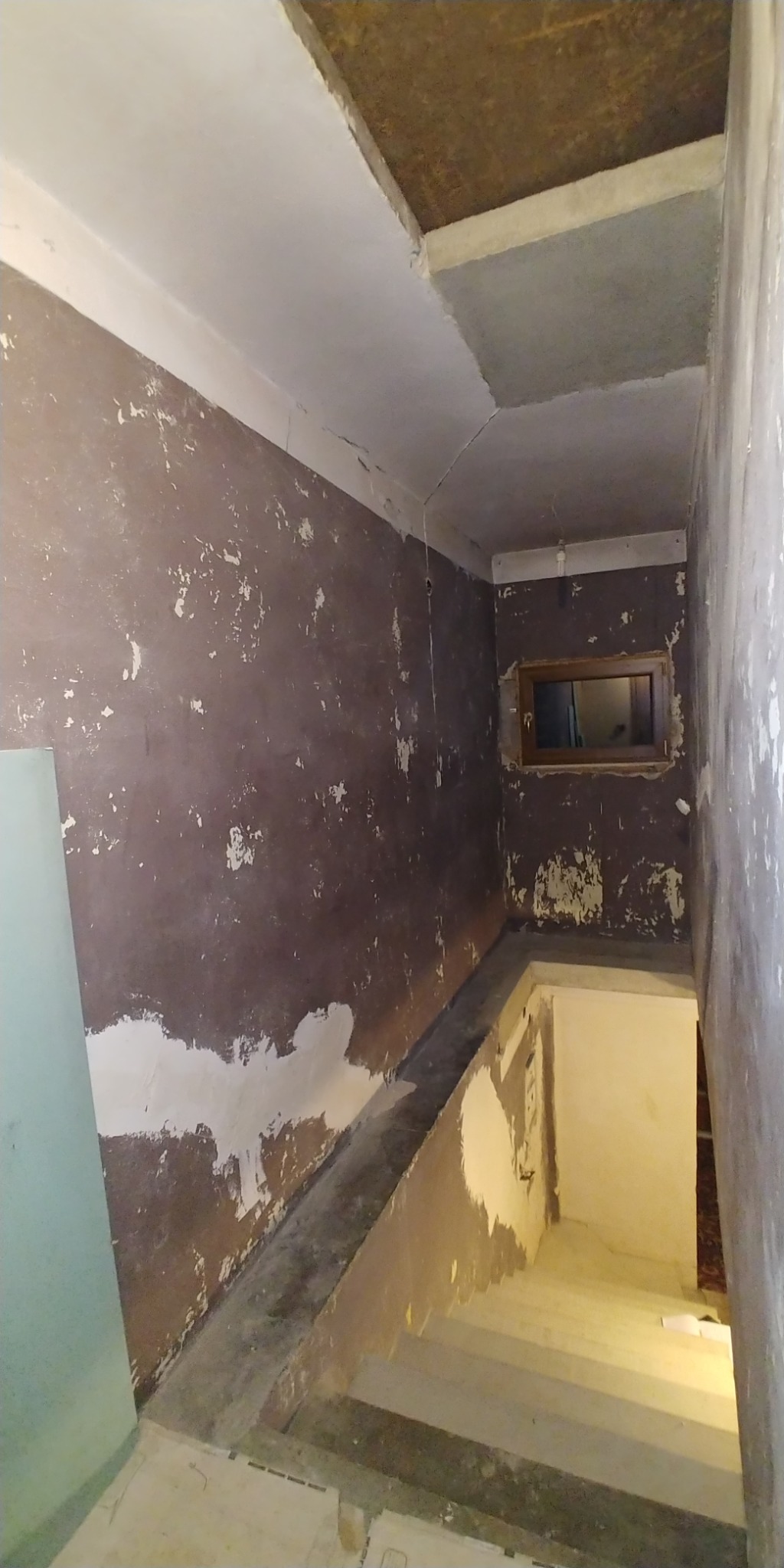 Klatka schodowa w trakcie prac przygotowawczych do remontu: ściany z resztkami starej farby i fragmentami gładzi, surowy sufit, schody z widocznymi śladami demontażu.