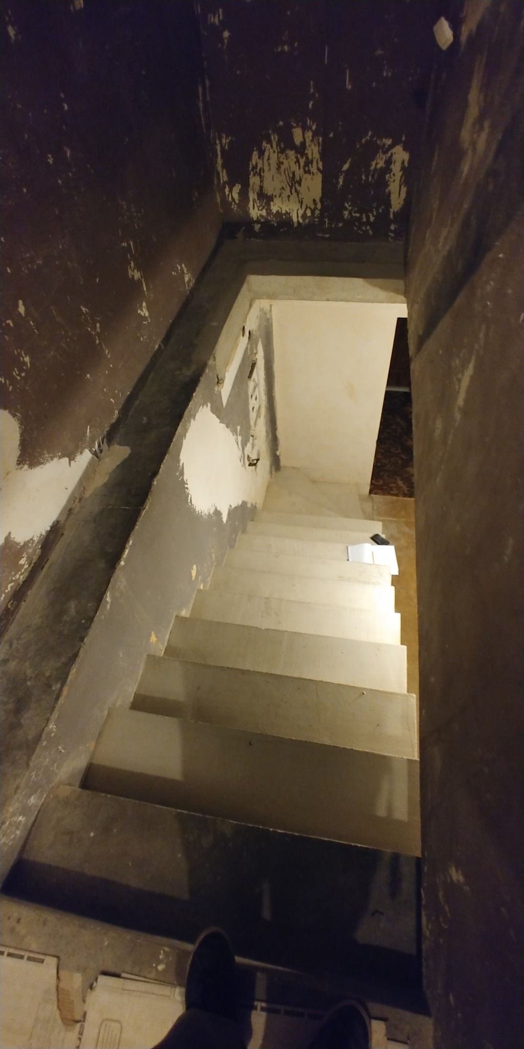 Klatka schodowa w trakcie remontu: ściany z resztkami starej farby i gładzi, surowe schody przygotowane do malowania, widoczne ślady po instalacjach elektrycznych.