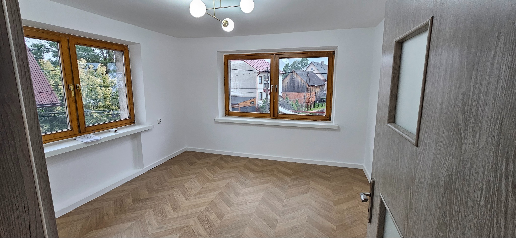 Wnętrze pokoju po remoncie: świeżo pomalowane białe ściany, parkiet ułożony we wzór jodełki, drewniane okna z widokiem na domy i zieleń, nowoczesna lampa sufitowa, szara drzwi z szybą.