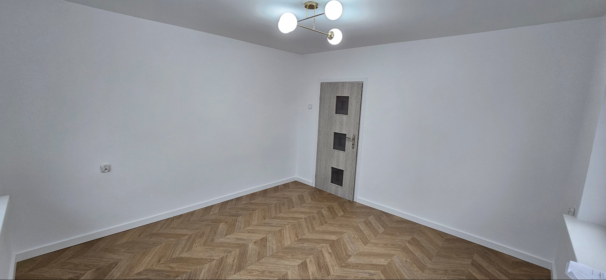 Pomalowane na biało ściany i ułożona parkietowa podłoga w pokoju z zamontowanymi drzwiami wewnętrznymi i nowoczesną lampą sufitową.