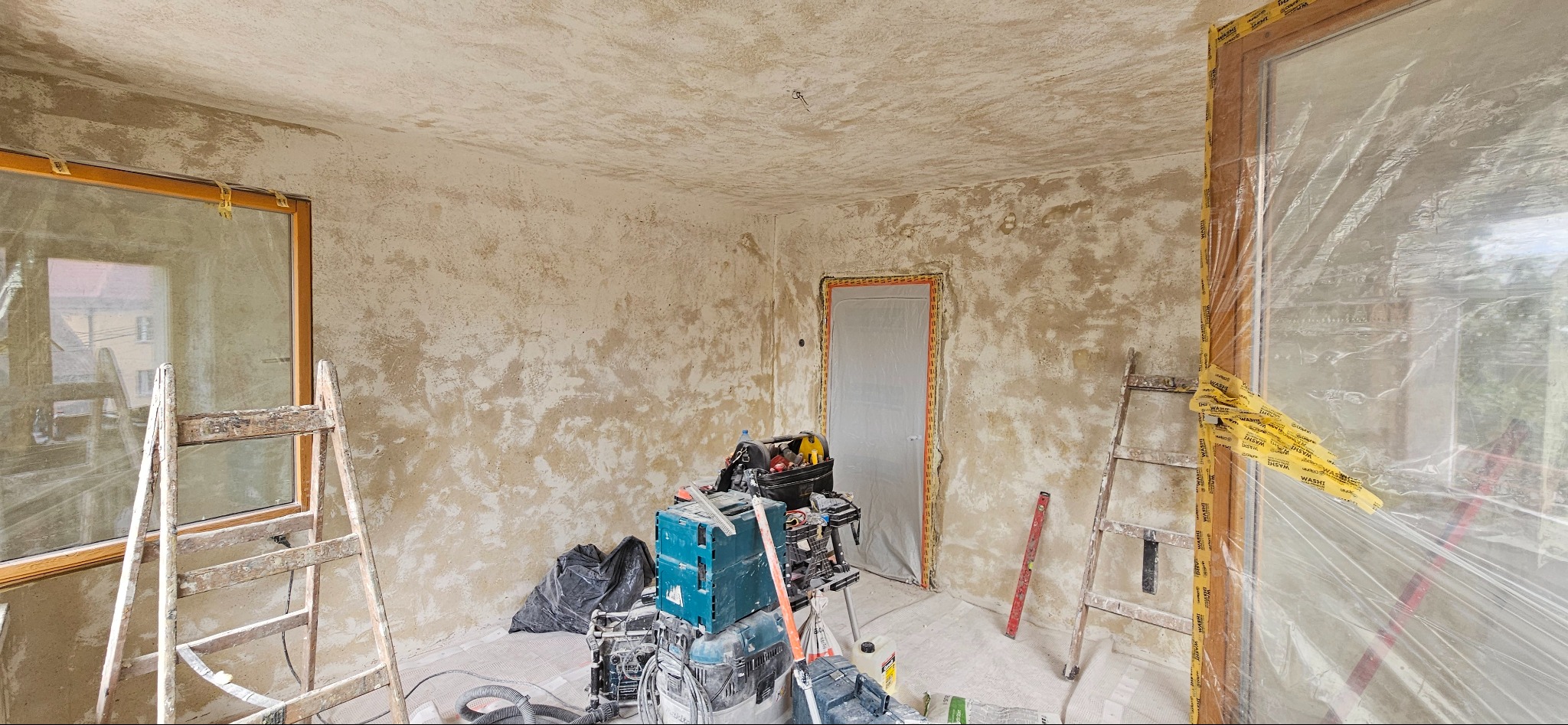 Wnętrze pokoju w trakcie prac wykończeniowych: ściany pokryte szpachlą, okna i drzwi zabezpieczone folią ochronną, drabiny malarskie i narzędzia budowlane widoczne na pierwszym planie.