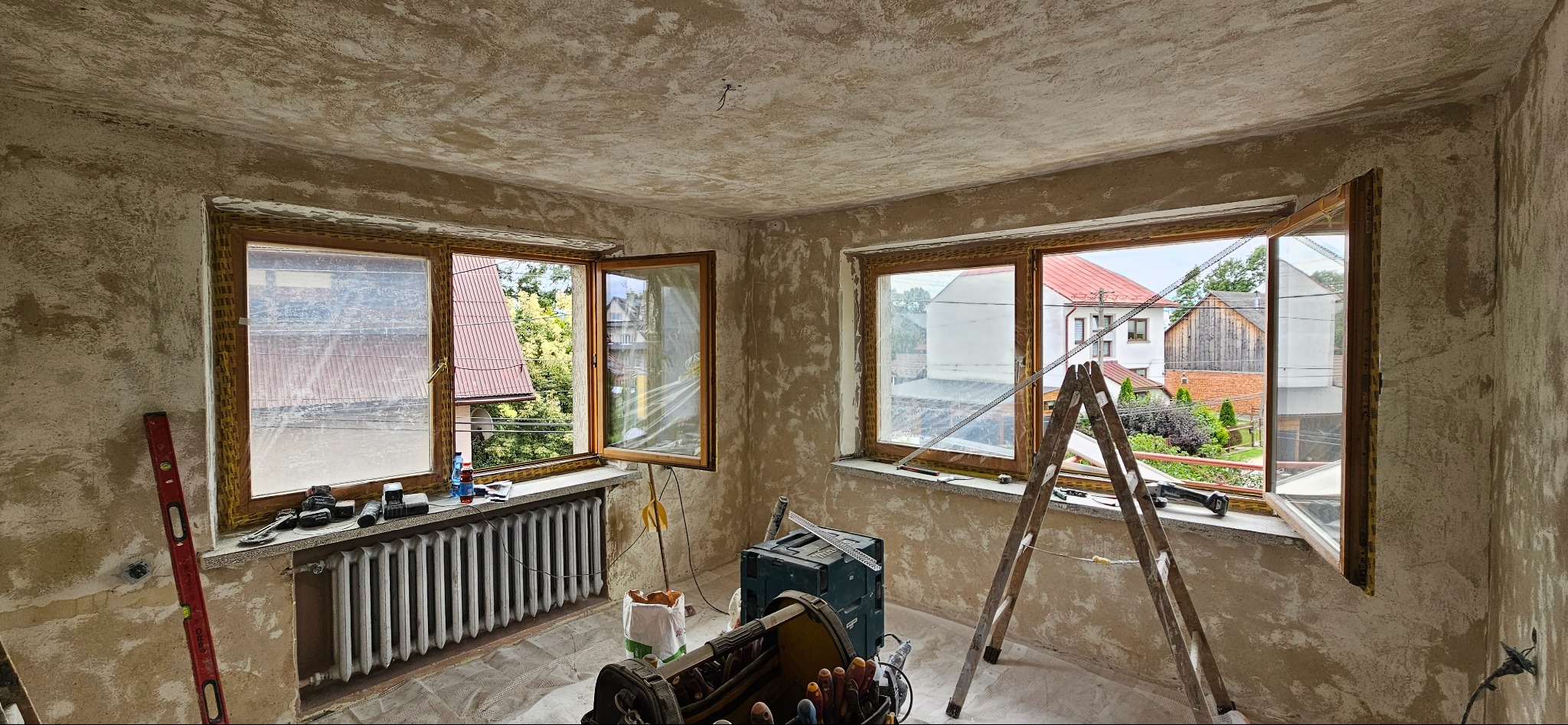 Pokój w trakcie prac wykończeniowych: ściany przygotowane pod gładzie, stare okna z drewnianą ramą, grzejnik, drabina i narzędzia budowlane.