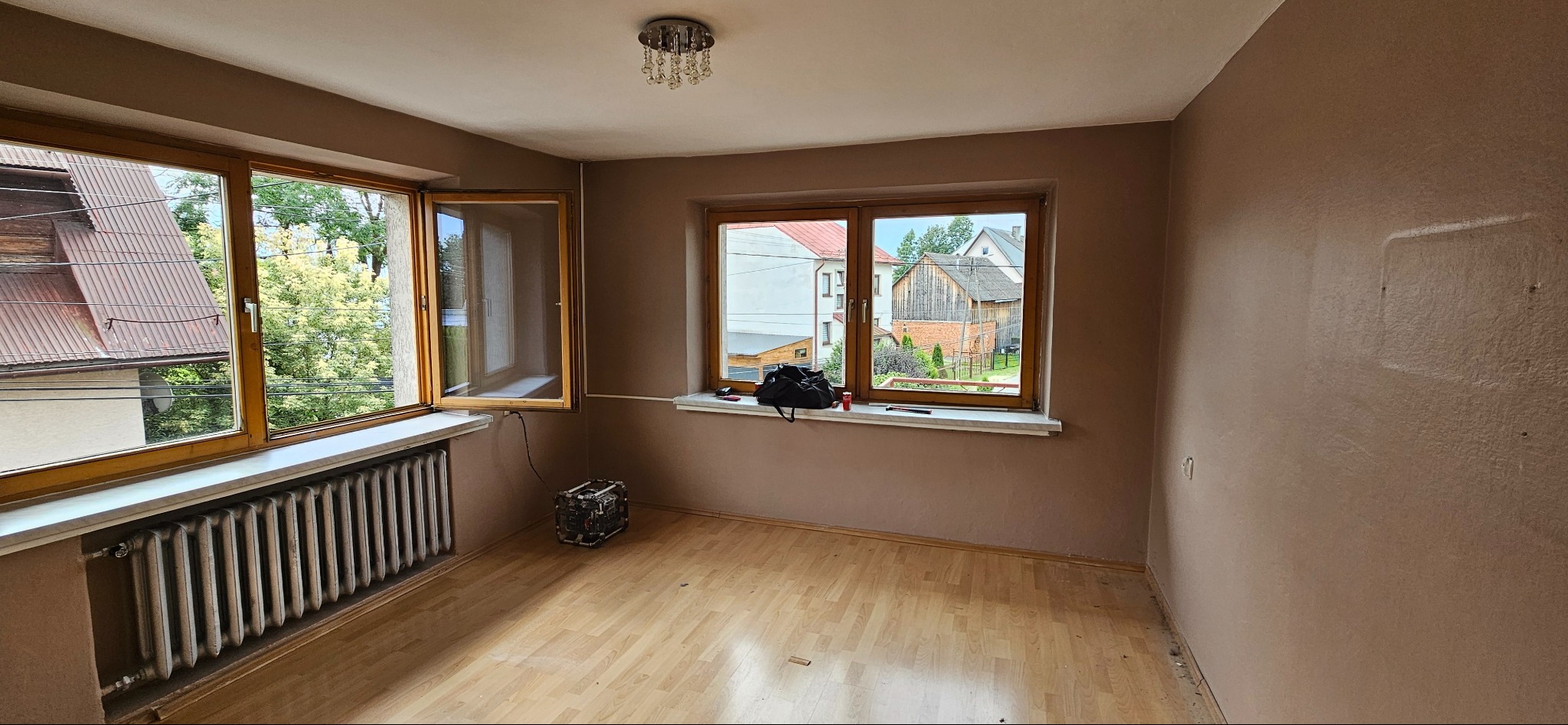 Pokój z dwoma oknami o drewnianych ramach, świeżo pomalowanymi ścianami w odcieniach beżu, parkietem na podłodze i starym grzejnikiem pod oknem po lewej stronie. Na parapecie pod oknem po prawej...