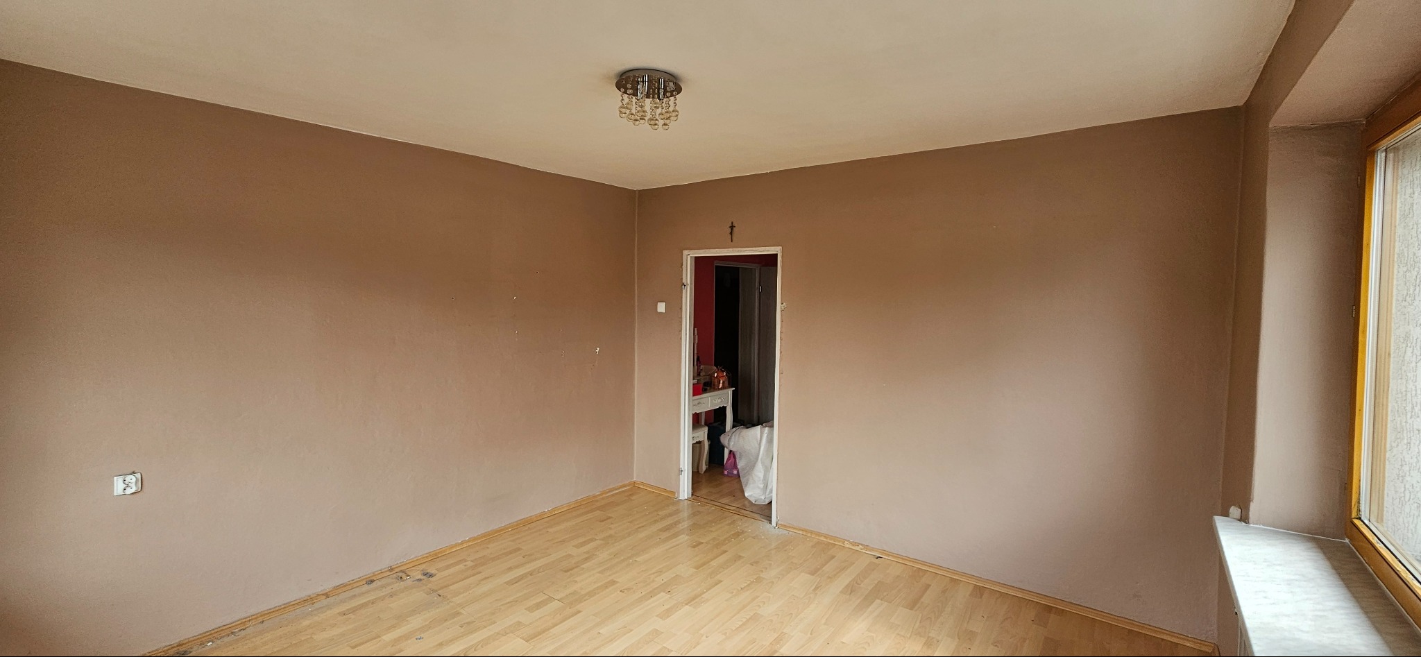 Pokój z brązowymi ścianami, drewnianą podłogą i widocznym oknem po prawej stronie. Na suficie wisi kryształowy żyrandol, a w tle widać otwarte drzwi do innego pomieszczenia.