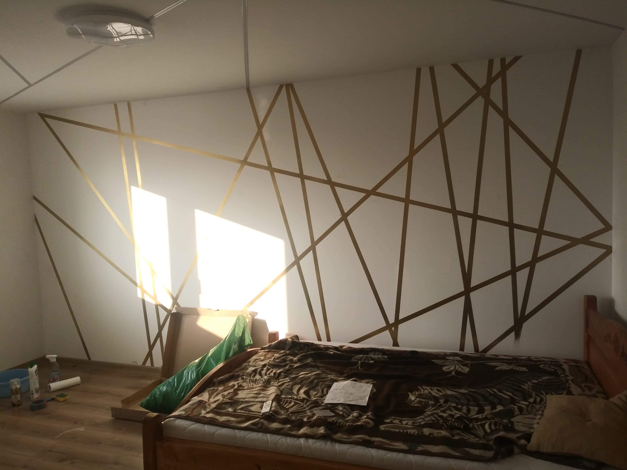 Ściana w pokoju z geometrycznym wzorem wykonanym z taśmy malarskiej, przygotowana do malowania, widoczne akcesoria malarskie i zabezpieczone meble.