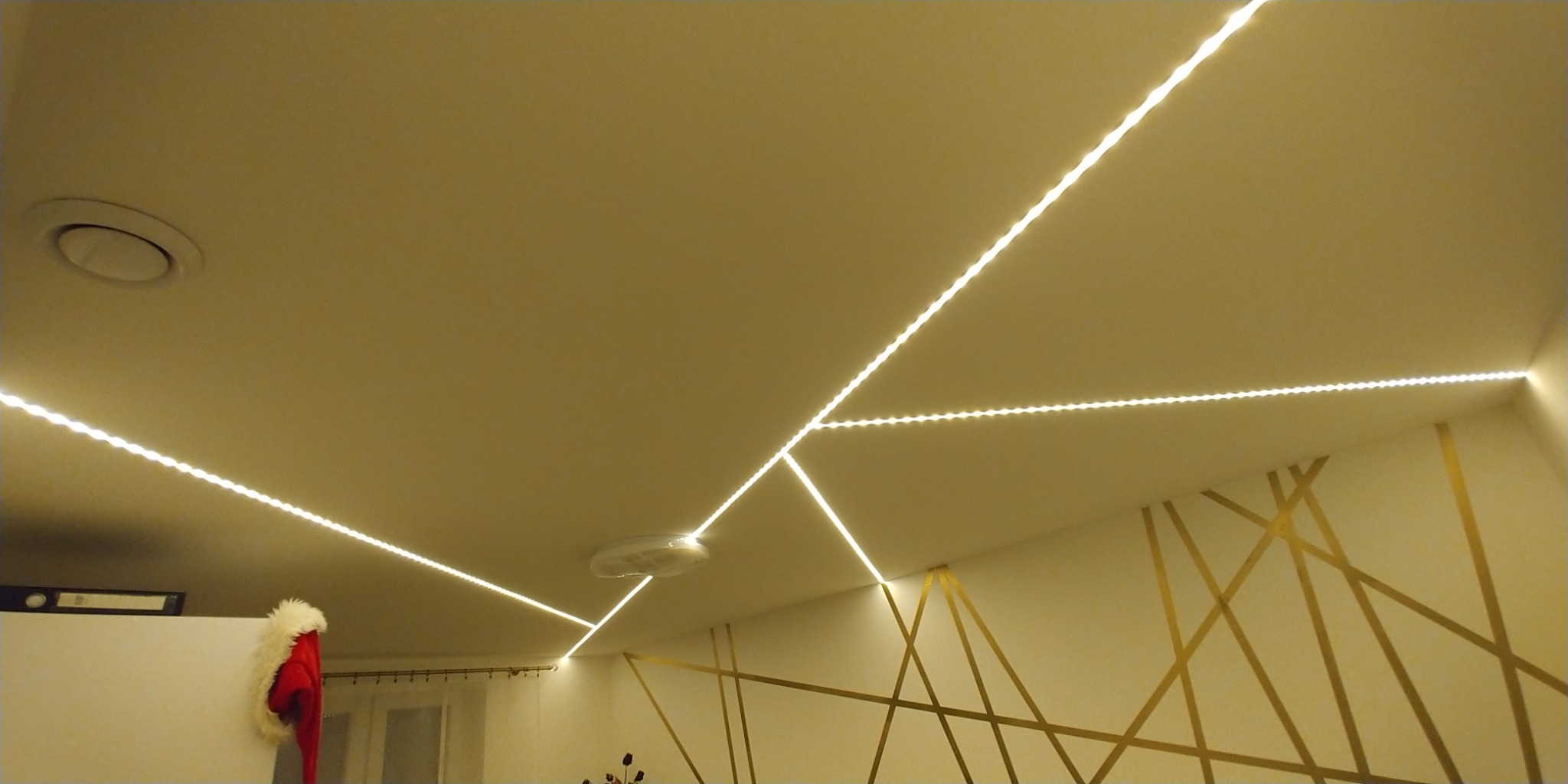 Biały sufit z zamontowanym oświetleniem LED w geometrycznym wzorze, uzupełniony złotymi geometrycznymi wzorami na ścianie i elementami dekoracyjnymi w tle.