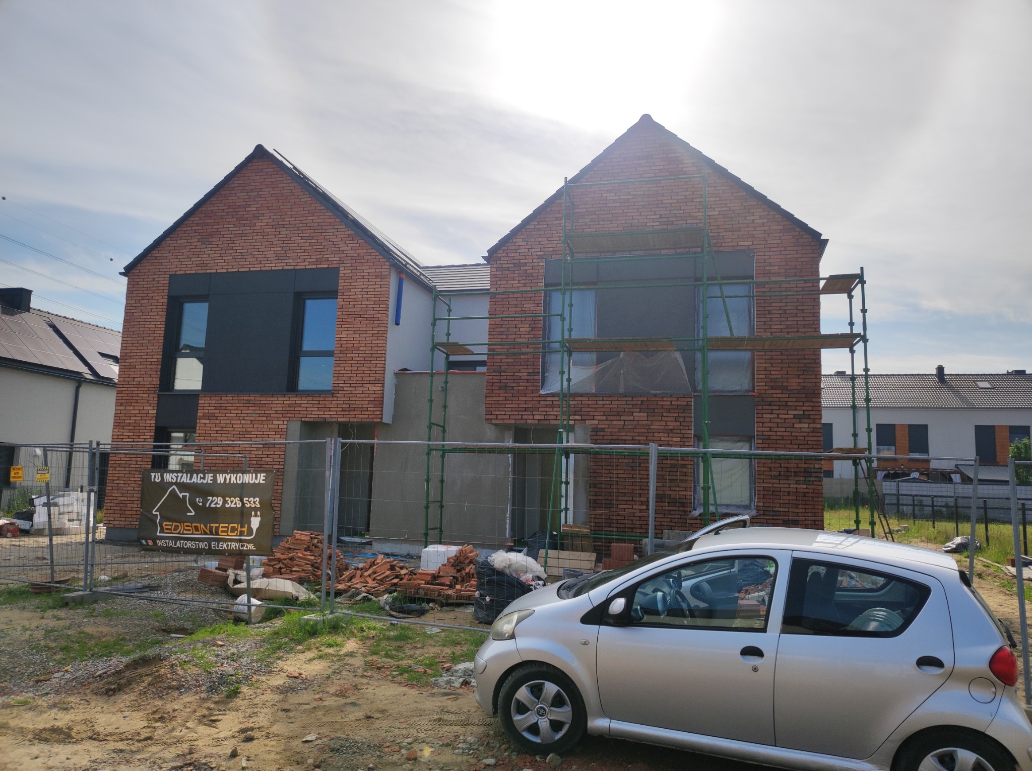 Nowo budowany dom z cegły, z widocznymi rusztowaniami i materiałami budowlanymi na posesji, częściowo ogrodzony, z zaparkowanym srebrnym samochodem z przodu.