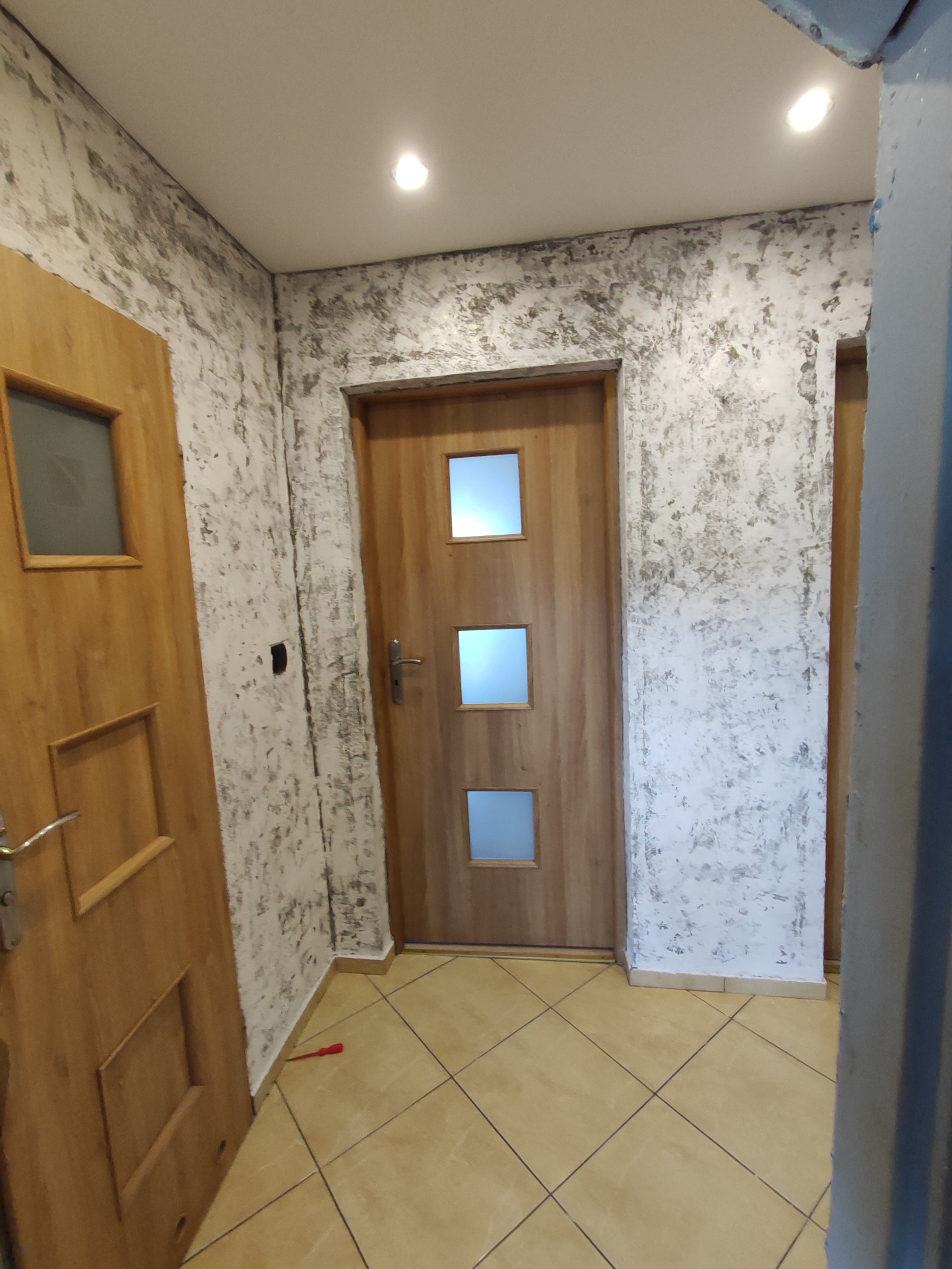 Korytarz z dwiema drewnianymi drzwiami, ściany z dekoracyjną szpachlą imitującą kamień, podłoga wyłożona płytkami, widoczny czerwony śrubokręt przy podłodze.