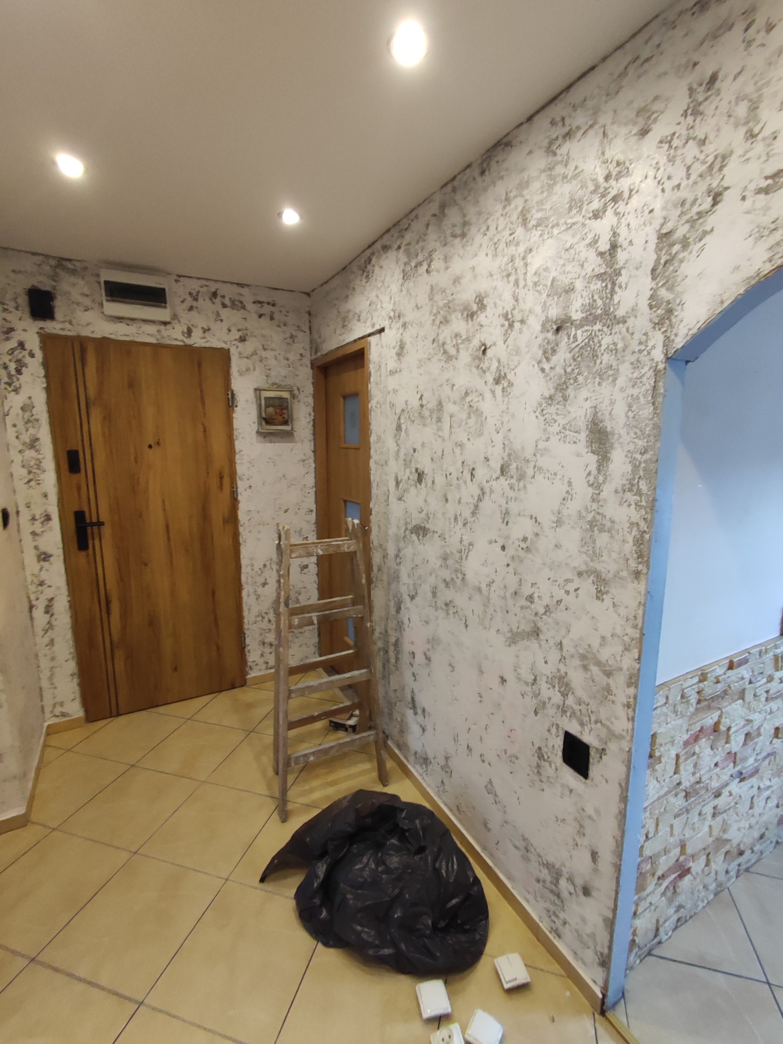 Korytarz w trakcie wykańczania z efektem dekoracyjnej, nieregularnej faktury na ścianach, widoczna drewniana drabina, zabezpieczona futryna drzwi i worek na śmieci z resztkami gipsu na podłodze.