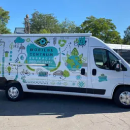 Biały van z grafiką edukacyjną o tematyce ekologicznej, prezentujący hasło 'Mobilne Centrum Edukacji Ekologicznej' w otoczeniu symboli recyklingu, energii odnawialnej i zrównoważonego transportu.