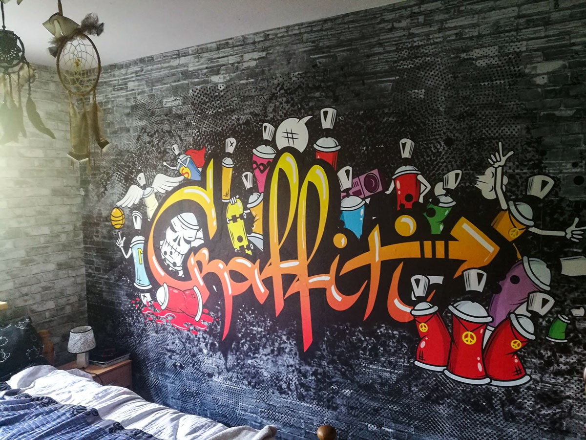 Ściana w pokoju pokryta grafitti z motywami czaszki, skrzydeł, puszek farby i napisu 'Graffiti' w stylu street art, widoczny fragment łóżka i łapacz snów.