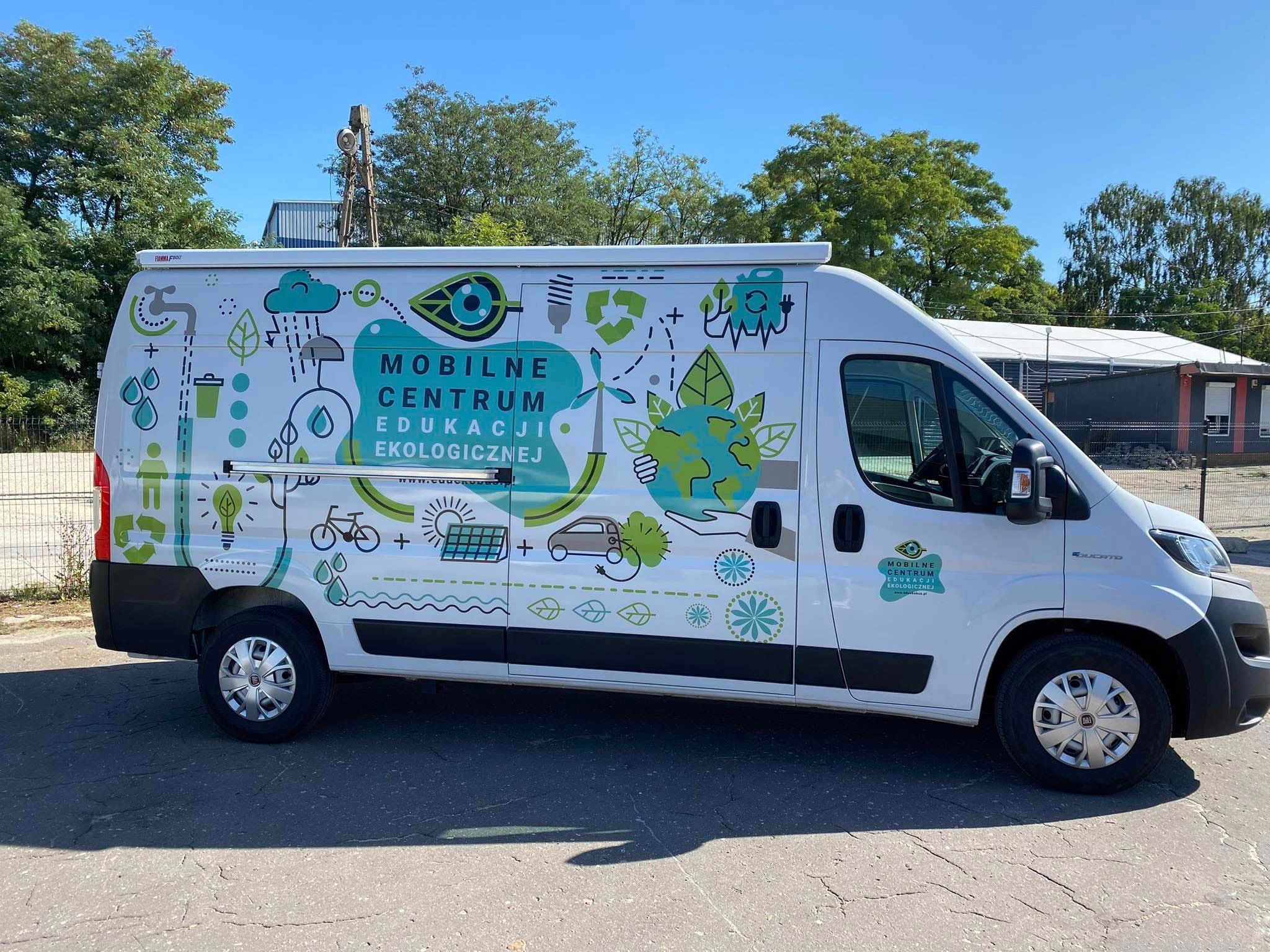 Biały van z grafiką edukacyjną o tematyce ekologicznej, prezentujący hasło 'Mobilne Centrum Edukacji Ekologicznej' w otoczeniu symboli recyklingu, energii odnawialnej i zrównoważonego transportu.