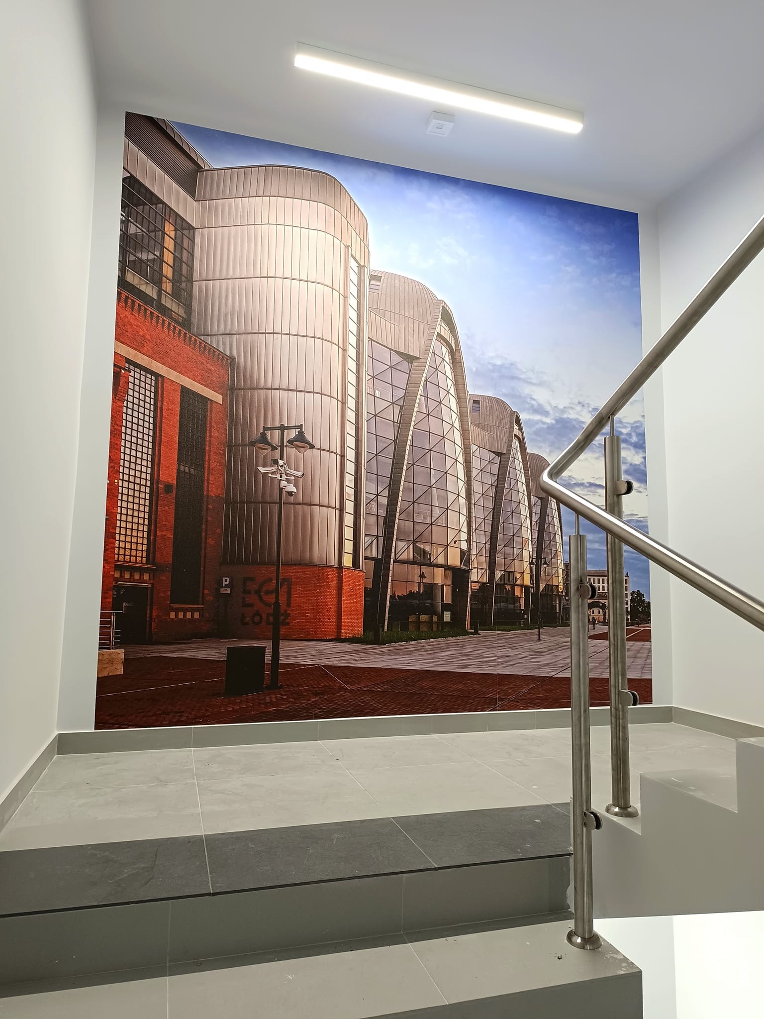 Fototapeta przedstawiająca budynek EC1 Łódź umieszczona na ścianie klatki schodowej, widoczna część schodów z szarych płytek oraz metalowa barierka.