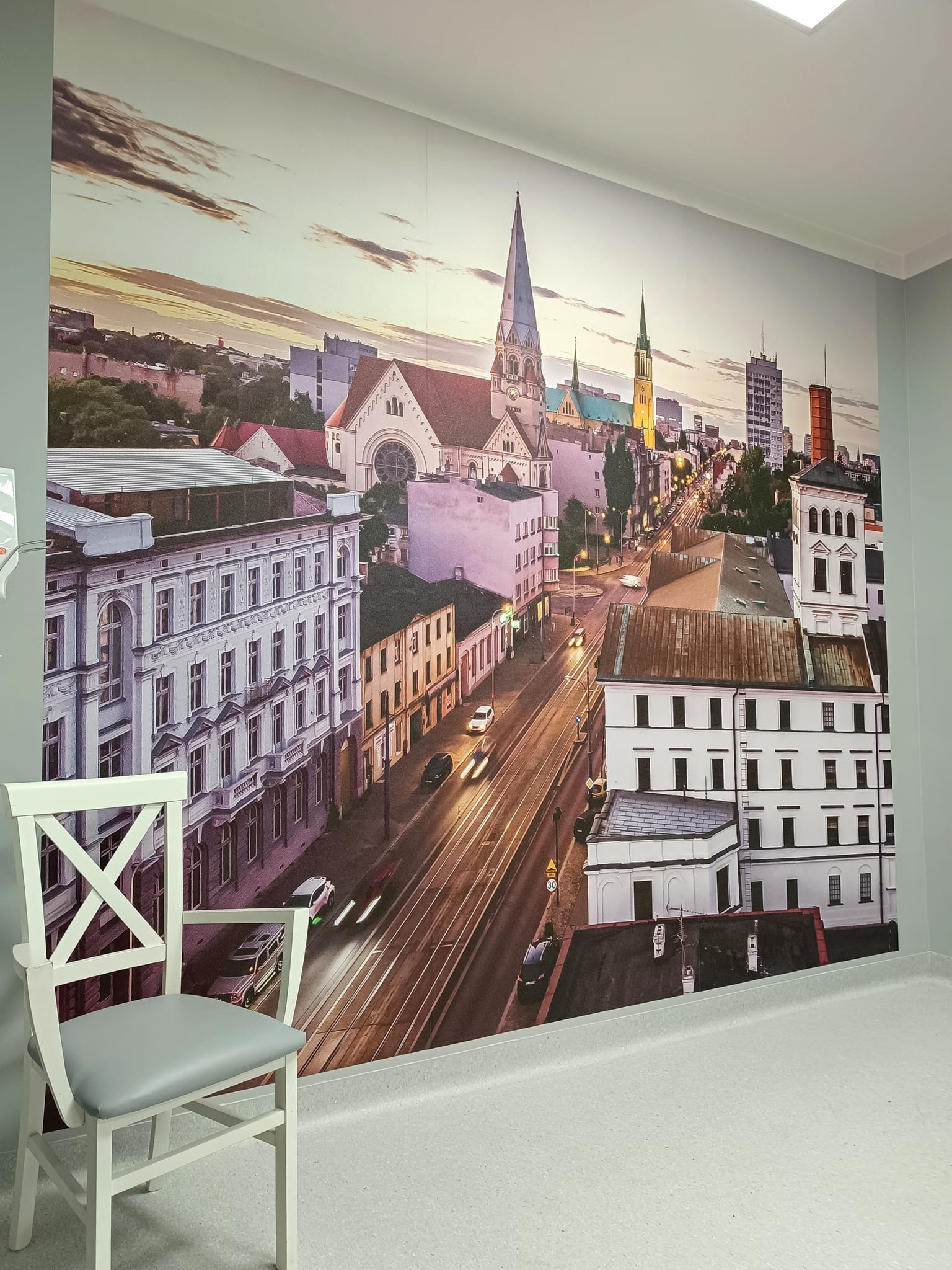 Wnętrze pokoju z fototapetą przedstawiającą panoramę miasta z kościołami i budynkami w ciepłych barwach zachodzącego słońca. Na pierwszym planie białe krzesło z szarym siedziskiem.