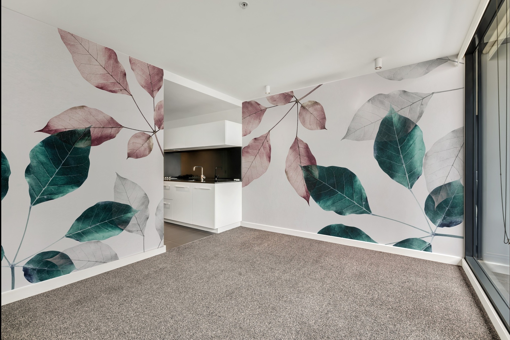 Wnętrze pokoju z fototapetą przedstawiającą liście w odcieniach zieleni, różu i szarości, widok na aneks kuchenny z białymi szafkami i czarnym blatem.