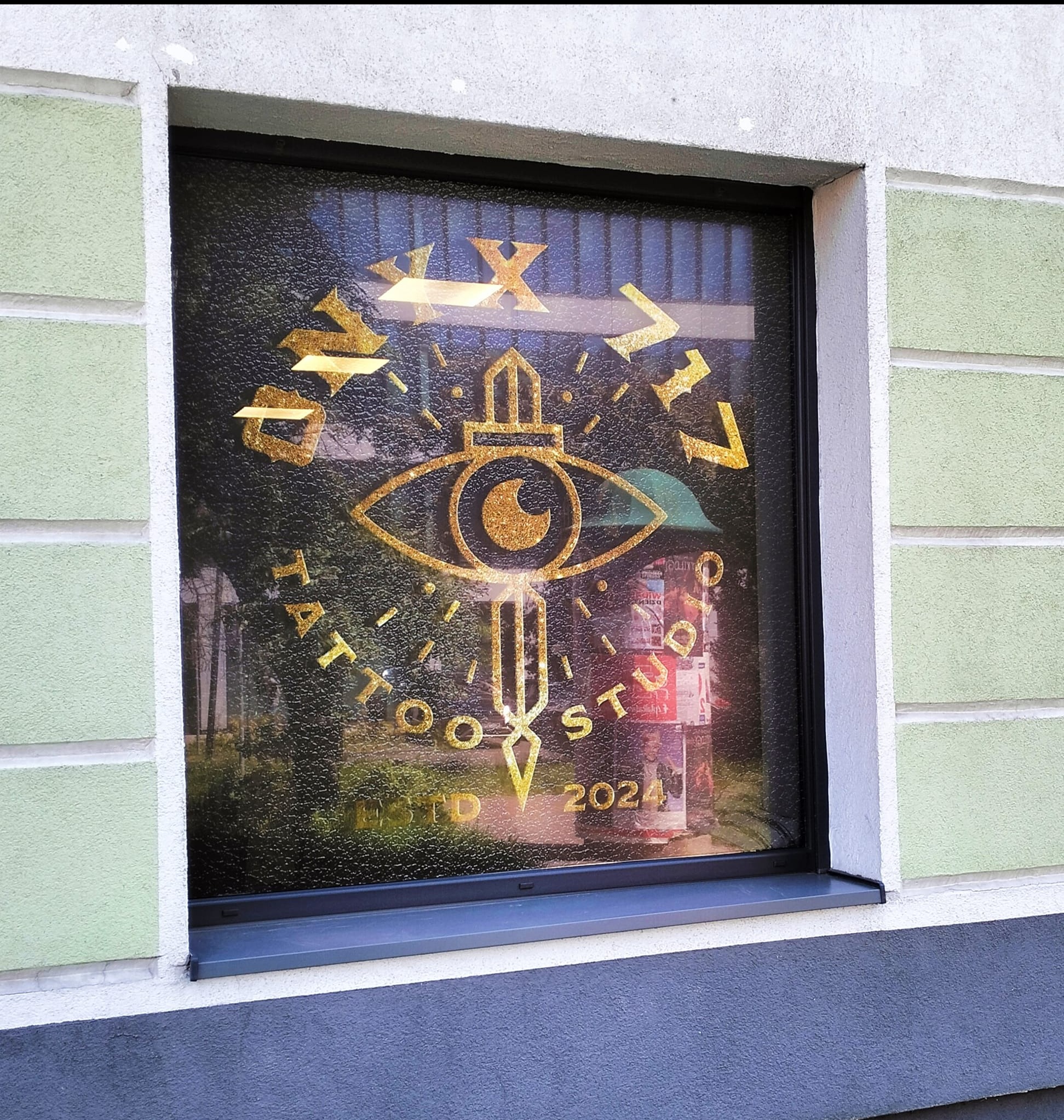 Złota grafika na oknie salonu tatuażu przedstawiająca oko z mieczem i napis 'TATTOO STUDIO ESTD 2024'.