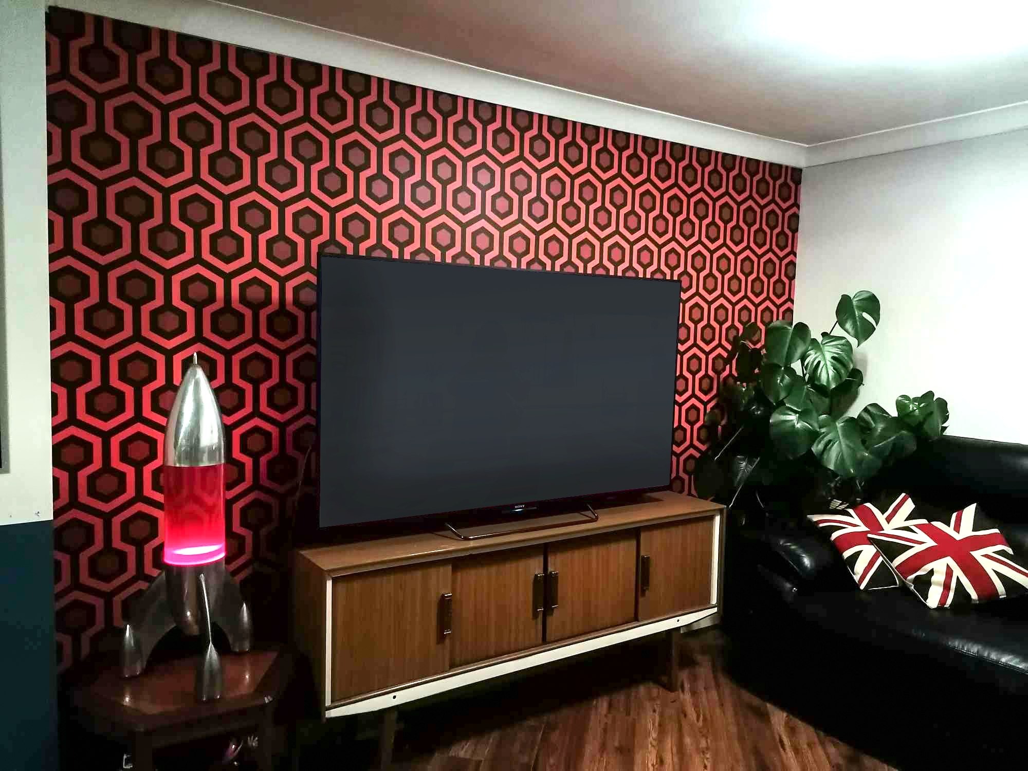 Salon z tapetą w geometryczny wzór w odcieniach czerwieni i brązu, telewizorem na szafce RTV w stylu vintage, lampą w kształcie rakiety i czarną skórzaną sofą z poduszkami w motywie flagi brytyjskiej.