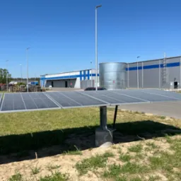 Rząd paneli słonecznych na metalowej konstrukcji przed budynkiem magazynowym z niebieskim pasem, w tle widoczny srebrny zbiornik i stosy palet.