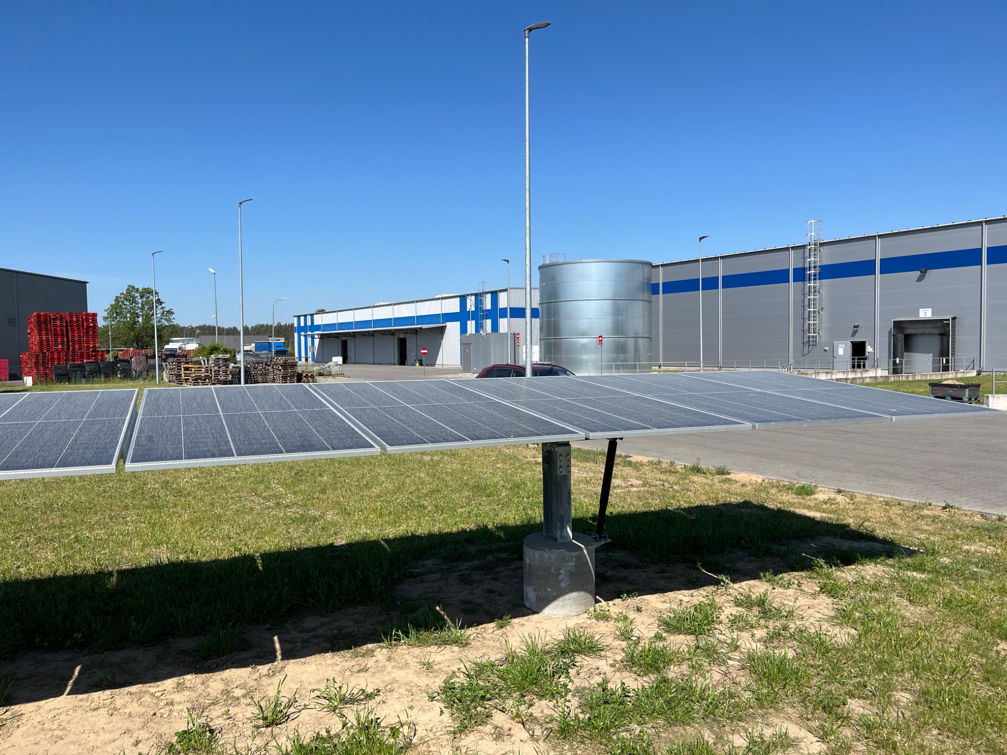 Rząd paneli słonecznych na metalowej konstrukcji przed budynkiem magazynowym z niebieskim pasem, w tle widoczny srebrny zbiornik i stosy palet.