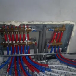 Rozdzielacz ogrzewania podłogowego z czerwono-niebieskimi rurami, zamontowany w metalowej skrzynce, uszczelnionej pianą montażową, widoczne maty izolacyjne.