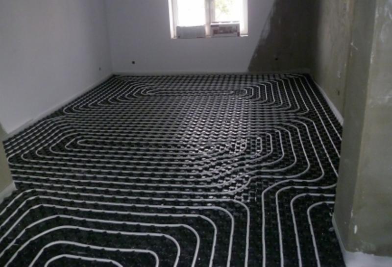 Ułożona instalacja ogrzewania podłogowego z białych rurek na czarnej macie w surowym pomieszczeniu z oknem. Widok z góry na całą powierzchnię.