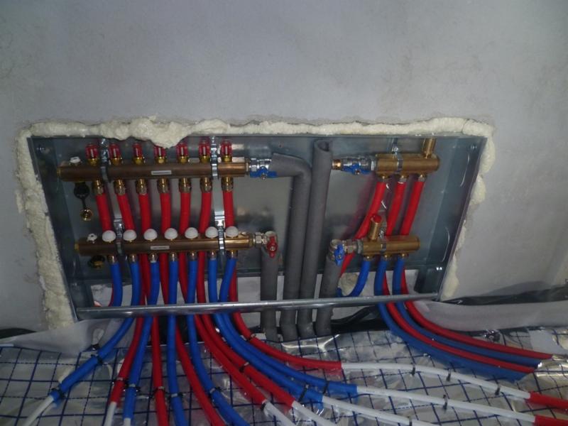 Rozdzielacz ogrzewania podłogowego z czerwono-niebieskimi rurami, zamontowany w metalowej skrzynce, uszczelnionej pianą montażową, widoczne maty izolacyjne.