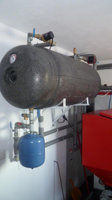 Szary, poziomy zbiornik akumulacyjny z manometrem, zaworami i niebieskim naczyniem przeponowym. Montaż na białych wspornikach, widoczne rury i fragment czerwonego kotła.