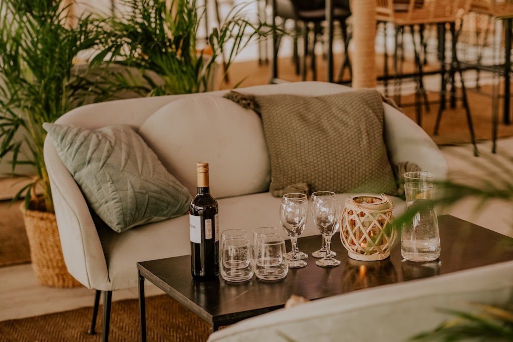 Stylowa aranżacja przestrzeni eventowej: butelka wina, różnorodne szklanki i świecznik na czarnym stoliku, miękka sofa z poduszkami i rośliny w tle, tworzące przytulną atmosferę.
