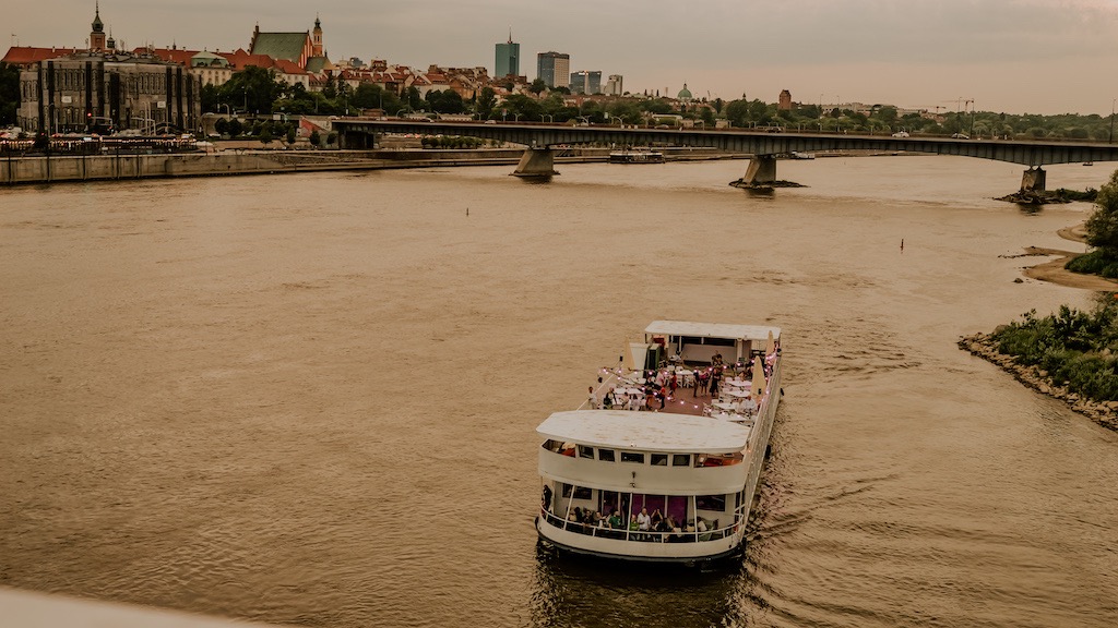 Statek wycieczkowy z oświetlonym pokładem i gośćmi na Wiśle, w tle panorama Warszawy i most Śląsko-Dąbrowski.