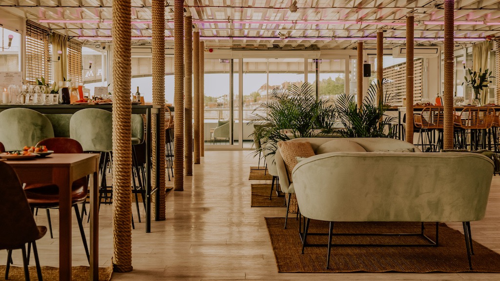 Wnętrze sali bankietowej na statku z dekoracyjnymi kolumnami owiniętymi liną, zielonymi sofami i krzesłami barowymi, przygotowane na przyjęcie.