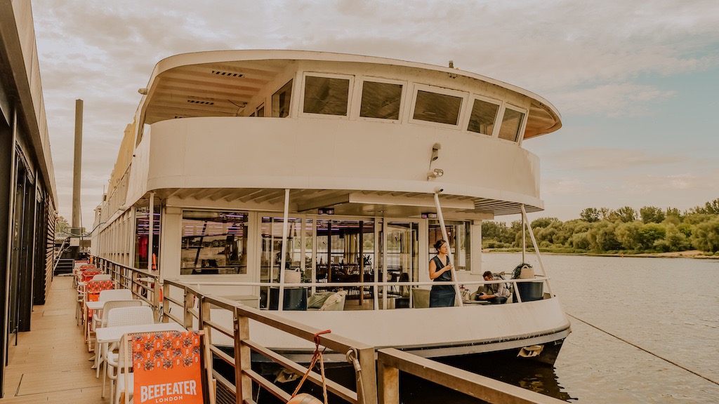Dwupoziomowy statek restauracyjny zacumowany przy brzegu rzeki, z zewnętrznym tarasem wyposażonym w stoliki i krzesła dla gości oraz reklamą marki Beefeater na barierkach.