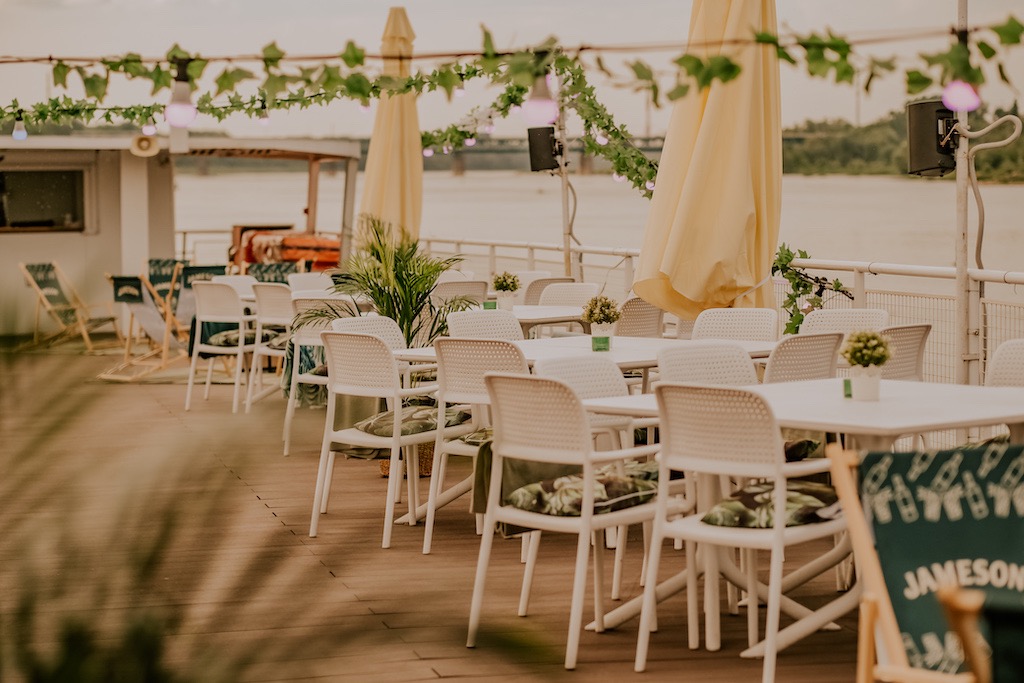 Ustawione białe krzesła wokół białych stolików na drewnianym pokładzie z widokiem na rzekę, dekoracje z zielonych liści i światełek, parasole przeciwsłoneczne, leżaki i tablica z napisem Jameson.