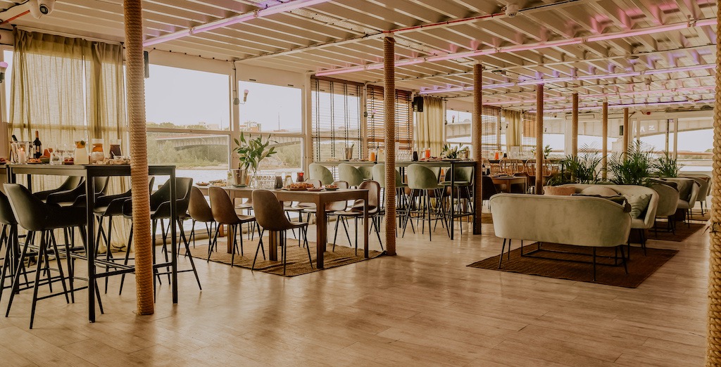 Elegancko urządzona przestrzeń eventowa na barce z widokiem na rzekę i most; wysokie stoliki barowe, stoły z nakryciami, sofy i rośliny doniczkowe tworzą przytulną atmosferę.