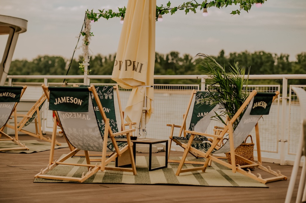 Zewnętrzna strefa relaksu z leżakami Jameson, stolikiem kawowym i parasolem na tle rzeki i zieleni, dekorowana girlandami świetlnymi.
