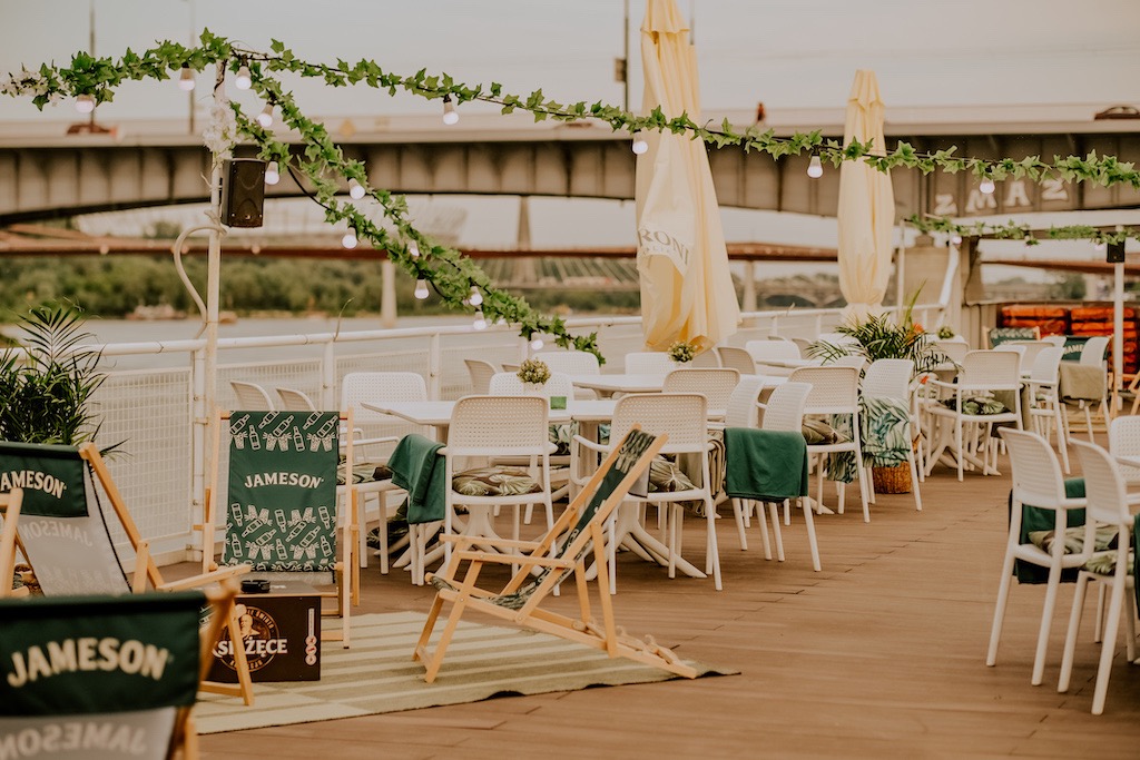 Dekoracja przestrzeni eventowej na zewnątrz z białymi krzesłami, stolikami, leżakami plażowymi z motywem Jameson i girlandami świetlnymi, widok na most w tle.