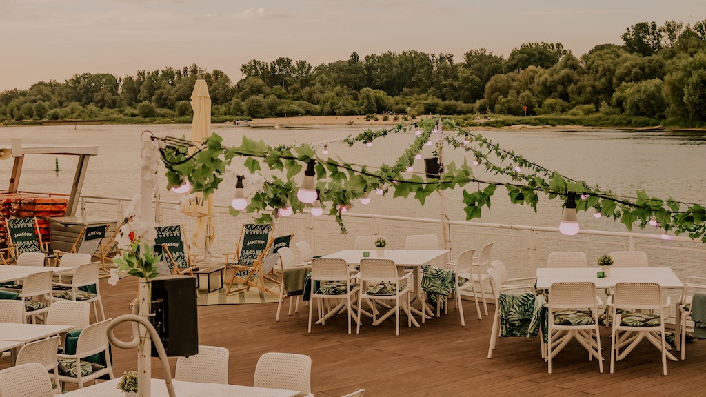 Dekoracje na barce: białe stoły i krzesła z zielonymi poduszkami, girlanda z żarówek i liści, widok na rzekę.