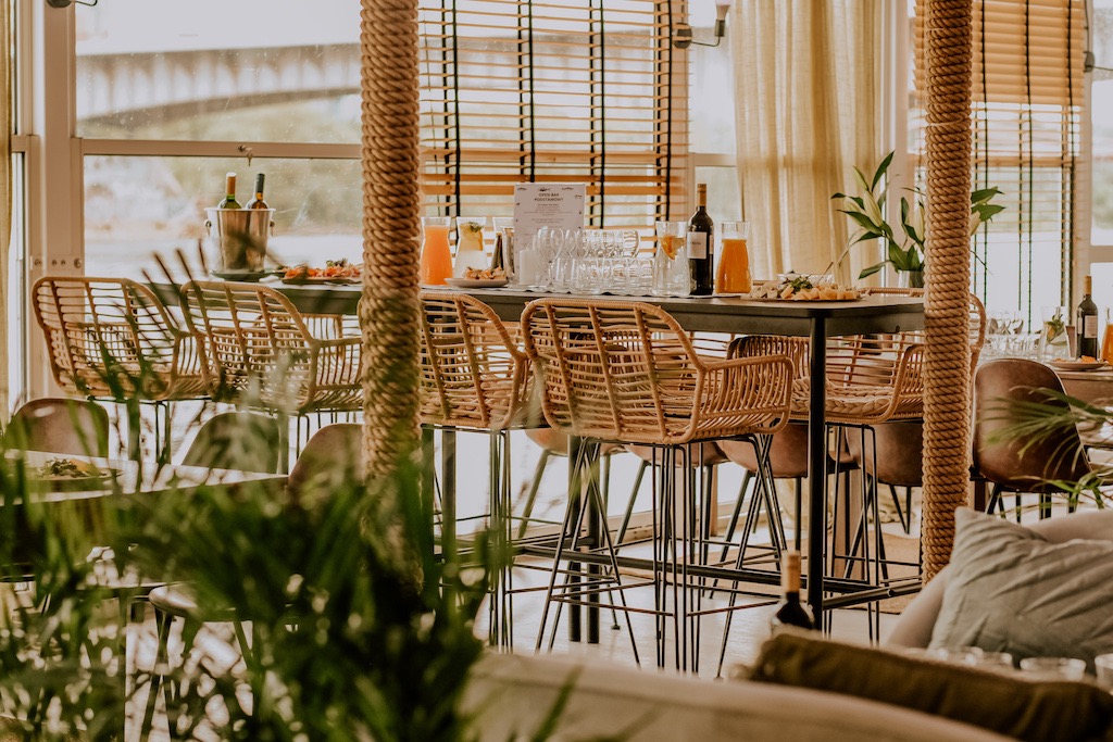 Uroczysta aranżacja wnętrza z wysokimi stołami, rattanowymi krzesłami barowymi, napojami w szklanych dzbankach i przekąskami, dekorowana liną jutową na filarach i zasłonami w tle.