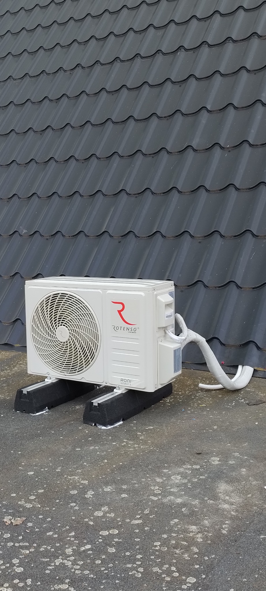 Biała jednostka zewnętrzna klimatyzacji marki Rotenso, model RONI, zamontowana na dachu pokrytym szarą blachodachówką, z izolowanymi rurami chłodniczymi.
