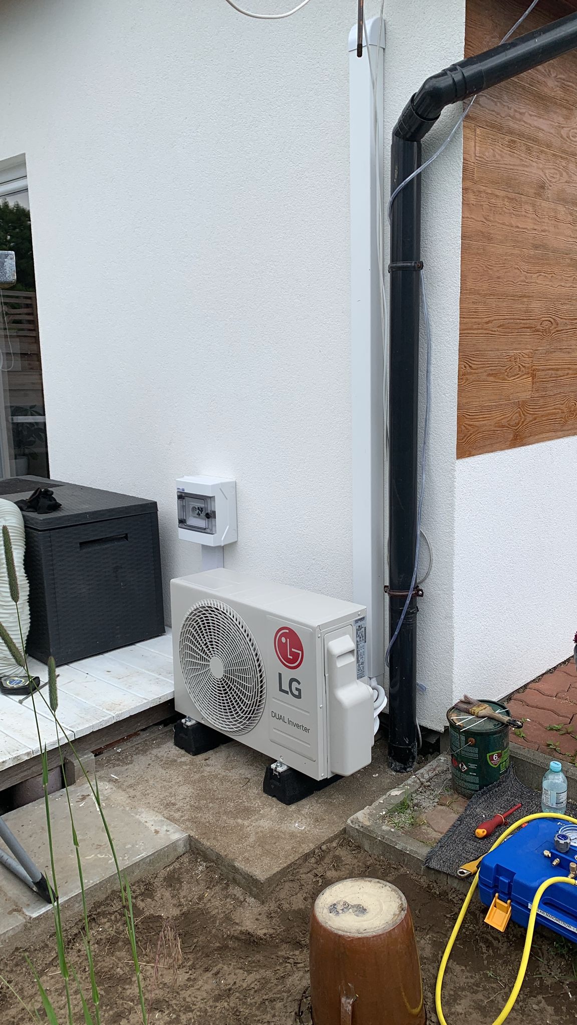 Zewnętrzna jednostka klimatyzacji LG Dual Inverter zamontowana na czarnych podstawkach przy białej ścianie budynku, obok rynny i skrzynki elektrycznej, z widocznymi narzędziami i butelką wody.
