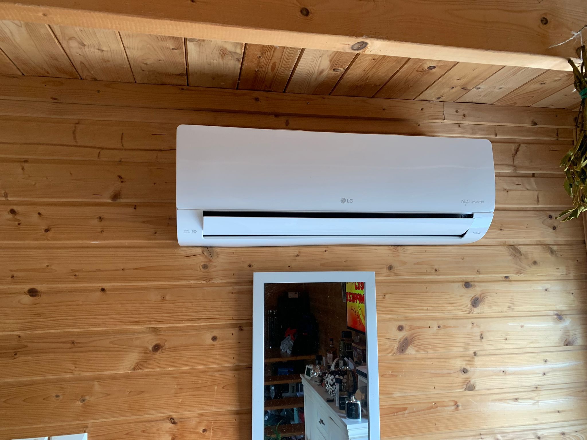 Biała jednostka wewnętrzna klimatyzacji LG Dual Inverter zamontowana na drewnianej ścianie z boazerii, pod drewnianym sufitem. W tle widoczne lustro z odbiciem wnętrza pokoju.