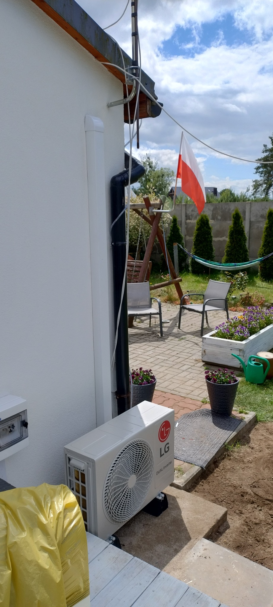 Zewnętrzna jednostka klimatyzacji LG Dual Inverter zamontowana przy ścianie domu z widocznym odpływem, w tle ogród z huśtawką i flagą Polski.