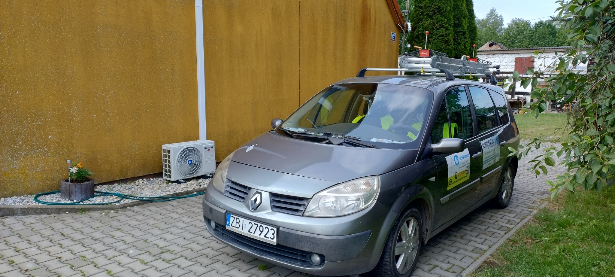 Szary samochód Renault z drabiną na dachu zaparkowany obok żółtej ściany budynku z zamontowaną jednostką zewnętrzną klimatyzacji Kaisai, na ścianie widoczny numer domu 12.